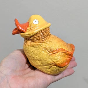 Könnte beinhalten: Gelb-orangefarbene Entenfigur mit strukturierter Oberfläche. Die Ente hat einen orangefarbenen Schnabel und ein einzelnes weißes Auge. Die Figur wird in einer Hand gehalten.