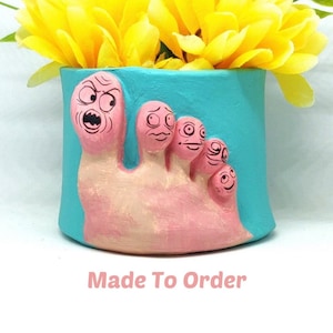 May include: A turquoise planter shaped like a foot with five toes, each with a different facial expression. The planter is filled with yellow flowers.
