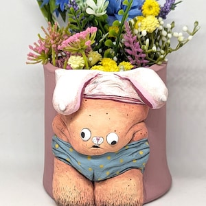 Könnte beinhalten: Ein rosafarbener Keramik-Pflanztopf in Form eines Comic-Kaninchens, das blaue und gelbe Unterwäsche mit Punkten trägt. Der Topf ist mit einem Strauß blauer, gelber und rosa Blumen gefüllt.