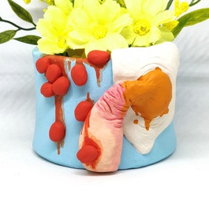May include: A blue ceramic planter with a white, orange and pink design. The planter has red paint splatters and a textured surface.