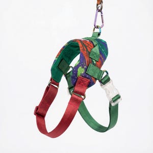 May include: A colorful dog harness with a green, red, and purple abstract pattern. The harness has red straps, green accents, and a clear plastic buckle. A purple carabiner clip is attached to the top.