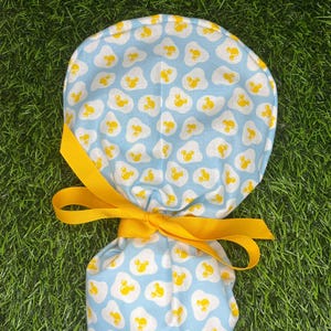 May include: A blue and yellow patterned scrub hat with a yellow ribbon tied around it. The pattern is of yellow fried eggs with a Mickey Mouse silhouette in the yolk.