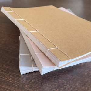 May include: Three notebooks stacked on top of each other, each with a different coloured cover. The top notebook has a brown cover, the middle one is pink, and the bottom one is grey. The pages are white, and the binding is white thread.