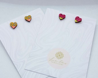 May include: Two pairs of heart-shaped stud earrings on white cards. One pair has a yellow top with blue dots and a pink bottom. The other pair has a pink top with red dots and a yellow bottom. The cards have a marble pattern.