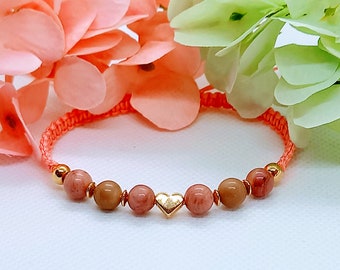 Puede incluir: Un brazalete rosa y naranja con un dije de corazón dorado y cuentas rosas y marrones.
