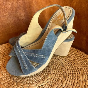 May include: Blue suede wedge sandals with a buckle closure and a criss-cross strap design. The sandals have a light brown platform and a light brown wedge heel.