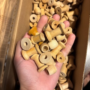 Pode incluir: Uma coleção de pequenas bobinas de madeira de cor clara com um centro oco, apresentando uma forma cilíndrica com extremidades alargadas. As bobinas são principalmente de madeira natural, com uma bobina exibindo um acabamento amarelo.