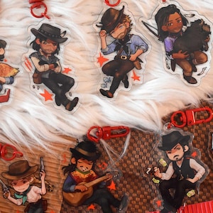 Puede incluir: Una colección de siete llaveros de acrílico transparente con ilustraciones estilo caricatura de personajes con atuendo del oeste. Cada llavero incluye un mosquetón de plástico rojo y muestra figuras con accesorios como sombreros, armas y guitarras.