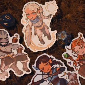 May include: Four illustrated stickers featuring fantasy characters. The stickers are on a brown fuzzy surface. The characters are a woman with blonde hair and a white outfit, a man with brown hair and a brown outfit, a woman with brown hair and a blue outfit, and a woman with brown hair and a green outfit.