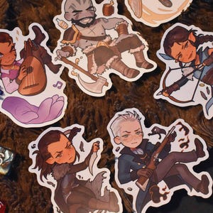 May include: A set of colourful stickers featuring various characters, including a warrior with an axe, a bard with a lute, a ranger with a bow and arrow, a rogue with a dagger, and a wizard with a staff. The stickers are on a brown, textured surface.