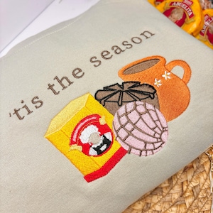 Può includere: Felpa beige ricamata con la frase "'tis the season" e immagini di una scatola gialla, un pasticcino marrone, una concha rosa e una tazza arancione. La scatola gialla ha un bordo rosso e un'immagine a cartoni animati.