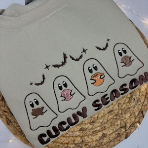 May include: A beige crewneck sweatshirt featuring an embroidered design of three white ghosts, each holding a different item, with small brown bats and stars above them. The text "CUCUY SEASON" is embroidered in brown letters at the bottom right.