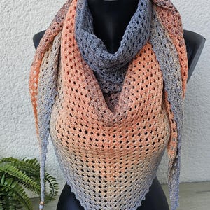 Könnte beinhalten: Ein gestrickter Dreiecksschal mit einem Farbverlauf von Pfirsich, Orange und Grau. Der Schal hat Fransen an den Enden und wird auf einer Schaufensterpuppe präsentiert.