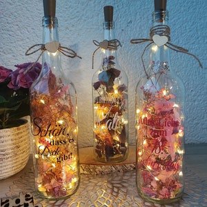 Könnte beinhalten: Drei Glasflaschen mit Korkstopfen und Bindfadenbögen, gefüllt mit getrockneten Blumen und Lichterketten. Die Flaschen haben handgeschriebene Botschaften: "Schon, dass es Dich gibt!" (German for "It's great that you exist!"), "Happy Birthday Kati" und "Alles Liebe zum 18. Geburtstag" (German for "All the best for your 18th birthday").