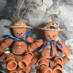 May include: Two terracotta pot dolls with straw hats and blue ribbon details. One doll is named "Trevor" and the other "Pam". The dolls have painted faces and are suspended by twine. A charming, handmade craft item.