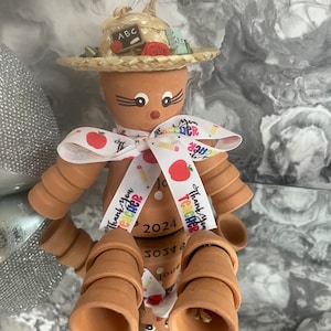 May include: A terracotta pot ornament shaped like a person wearing a straw hat with a red flower and the letters "ABC" on a small chalkboard. The ornament has a white ribbon with red apples and the text "Thank you Teacher 2024".