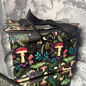 May include: Black insulated lunch bag featuring a vibrant mushroom and moth design. The bag has a black zip, handle, and trim. The pattern includes red, pink, and white mushrooms, green moths, and various floral elements.