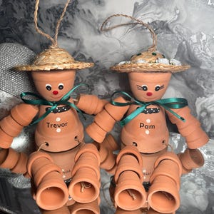May include: Two terracotta pot dolls with straw hats and green ribbon bows. One doll is labelled "Trevor" and the other "Pam". The dolls have painted faces and are connected with string, ideal for home decor or as a unique gift.