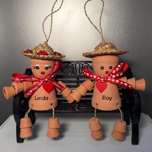 May include: Two terracotta pot figures, "Linda" and "Roy", sit on a black bench. They wear straw hats, red heart accents, and red ribbon. The figures have pot arms and legs, and are connected by holding hands. A Valentine's Day theme.