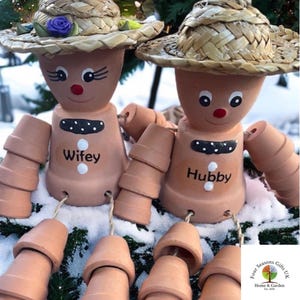 May include: A pair of terracotta planter people with straw hats, painted faces, and the words "Wifey" and "Hubby" on their bodies. They are standing in the snow with their arms and legs made of stacked terracotta pots.