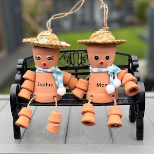 May include: Two terracotta pot figures, each wearing a straw hat and scarf, sit on a black bench. One figure is labelled "Sophie" and the other "Max". The figures have painted faces and dangling legs, connected by string.