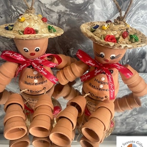 May include: Two terracotta pot people with straw hats and red ribbons. The pot people are holding signs that say "Happy Retirement" with names Brian and Diane.