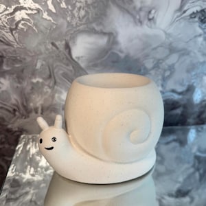 May include: A cream-coloured snail-shaped planter with a smiling face and small antennae. The planter has a round body with a spiral design and a large opening for plants. The planter is made of a speckled material.