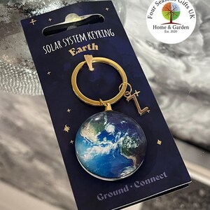 May include: Gold-coloured keyring with a blue and green globe charm. The charm has a white background with a detailed image of Earth. The keyring is labelled "Solar System Keyring Earth" and "Ground Connect".