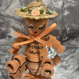 May include: A terracotta pot figurine of a person wearing a straw hat with a pink flower and a peach-colored ribbon. The figurine has the words "Happy Birthday Mum" written on it.