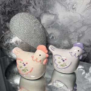 May include: A pair of ceramic hen-shaped salt and pepper shakers. One is light pink with a peach comb and floral details, the other white with a lavender comb and floral details. Both have a glossy finish and are reflected on a mirrored surface.