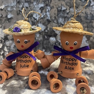 May include: Two terracotta pot people with straw hats and purple bows. The pots are decorated with the text "Happy Birthday" and the names "Julie" and "Alex".