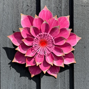 Puede incluir: Una gran flor de metal rosa con m&uacute;ltiples capas de p&eacute;talos y un centro rojo. Los bordes de los p&eacute;talos tienen un borde dorado. La flor est&aacute; montada sobre una superficie de madera gris oscuro, creando un contraste llamativo.