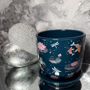 May include: A teal ceramic plant pot with a matching saucer. The pot is decorated with a pond scene featuring pink lotus flowers, koi carp, dragonflies, and a frog. The pot has a glossy finish.