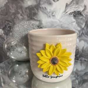 May include: A cream ceramic plant pot with a textured surface and a bright yellow sunflower applique. The pot has the phrase "Hello sunshine" written on the base. A decorative silver sphere is in the background.