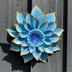 Puede incluir: Una gran flor de metal azul con ribetes dorados, montada sobre una superficie de madera gris. La flor tiene m&uacute;ltiples capas de p&eacute;talos y un centro detallado. La sombra de la flor es visible en la superficie de madera.