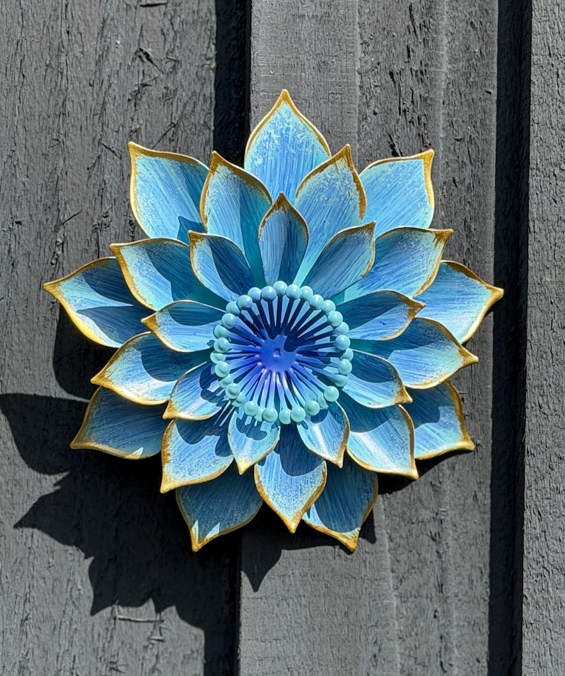 Puede incluir: Una gran flor de metal azul con ribetes dorados, montada sobre una superficie de madera gris. La flor tiene m&uacute;ltiples capas de p&eacute;talos y un centro detallado. La sombra de la flor es visible en la superficie de madera.