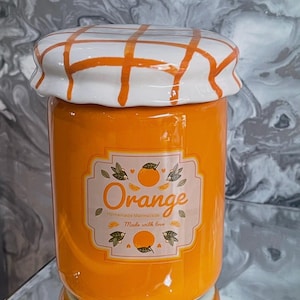 May include: A bright orange ceramic jar with a white lid and orange plaid design. The jar has a label that reads "Orange Homemade Marmalade Made with love."