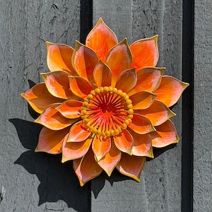 Puede incluir: Escultura floral met&aacute;lica vibrante con tonos naranja, amarillo y rosa. Los p&eacute;talos tienen un efecto degradado y el centro presenta un dise&ntilde;o texturizado detallado. Montada sobre una superficie de madera gris.