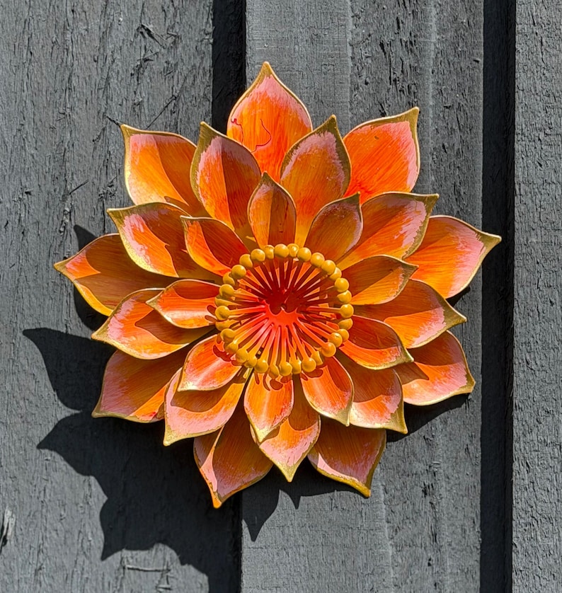 Puede incluir: Escultura floral met&aacute;lica vibrante con tonos naranja, amarillo y rosa. Los p&eacute;talos tienen un efecto degradado y el centro presenta un dise&ntilde;o texturizado detallado. Montada sobre una superficie de madera gris.
