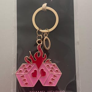 May include: Pink enamel keyring featuring two dice with heart symbols, set against red flames. The keyring has a gold-coloured ring and a charm with the number zero. The packaging reads "a roll of the dice" and "ENAMEL KEYRING."