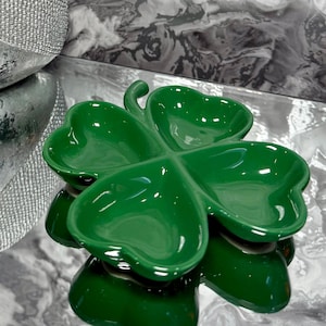 May include: A glossy, green ceramic clover-shaped dish. The dish has four heart-shaped sections and a curved stem. The dish is sitting on a reflective surface, with a blurred background.