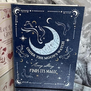 May include: A navy blue book with a celestial design. The cover features a crescent moon with a face, stars, and the text "UNDER THE MOON'S WATCH" and "EVERY STORY FINDS ITS MAGIC". The book is part of a series.