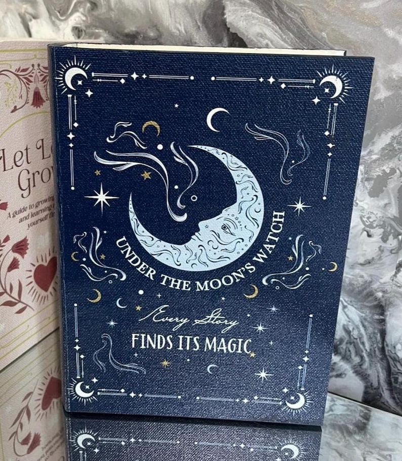 May include: A navy blue book with a celestial design. The cover features a crescent moon with a face, stars, and the text "UNDER THE MOON'S WATCH" and "EVERY STORY FINDS ITS MAGIC". The book is part of a series.