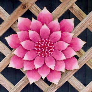 Puede incluir: Una gran flor de metal rosa con un dise&ntilde;o detallado. La flor tiene m&uacute;ltiples capas de p&eacute;talos con un borde rosa m&aacute;s claro y un centro texturizado. Est&aacute; montada sobre un fondo de celos&iacute;a de madera, creando un acento decorativo para el jard&iacute;n.