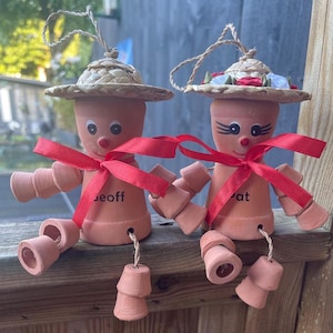 May include: Two terracotta pot figures with straw hats and red ribbon bows. The figures have painted faces and are seated. One figure has the name "Geoff" and the other has the name "Pat" written on them. They are hanging ornaments.