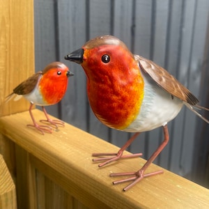 May include: Two metal robin figurines, one larger than the other, with red breasts and white bellies. The larger robin is perched on a wooden surface, while the smaller robin is standing on the surface behind it.