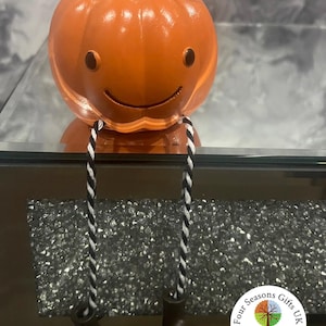 May include: Orange pumpkin ornament with a smiling face, brown eyes, and a green stem. It has black and white striped legs and brown boots. A decorative item for the home.