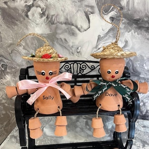 May include: Two terracotta pot figures, "Sally" and "Dave", sit on a black bench. Each figure has a straw hat, painted face, and ribbon. "Sally" has a pink bow, "Dave" a green one. The figures' arms and legs are made of small pots.