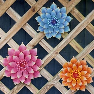 Puede incluir: Tres grandes decoraciones florales coloridas montadas en un enrejado de madera. Las flores son en tonos de azul, rosa y naranja, cada una con dise&ntilde;os detallados de p&eacute;talos y centros contrastantes. El enrejado es de madera marr&oacute;n claro.