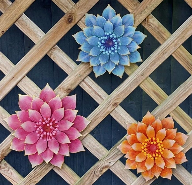 Puede incluir: Tres grandes decoraciones florales coloridas montadas en un enrejado de madera. Las flores son en tonos de azul, rosa y naranja, cada una con dise&ntilde;os detallados de p&eacute;talos y centros contrastantes. El enrejado es de madera marr&oacute;n claro.